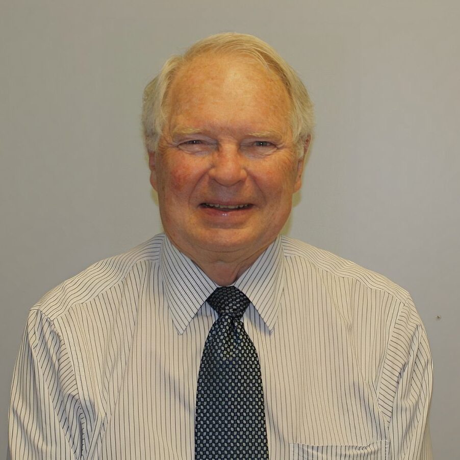 Older man with white hair wearing a striped shirt and polka dot tie, posing in front of a neutral background.